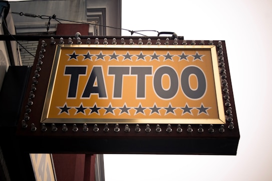 A rectangular sign with marquee lights features the word 'TATTOO' prominently displayed in bold, capital letters. The background is yellow, decorated with black stars above and below the text. The sign appears to be mounted on a building exterior.