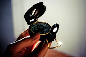 A hand holds an antique-style, black and gold compass. The compass is open with its lid flipped up, revealing a decorative face with intricate markings. Subdued lighting creates a warm, slightly shadowed atmosphere.