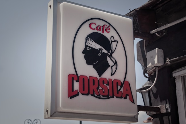 A rectangular sign displays the silhouette of a person's head with a bandana over the forehead, accompanied by the text 'Café Corsica'. The silhouette is in a bold black, while the text is a deep red and uppercase. The background of the sign is white, and the image is attached to a dark wall with visible wires.