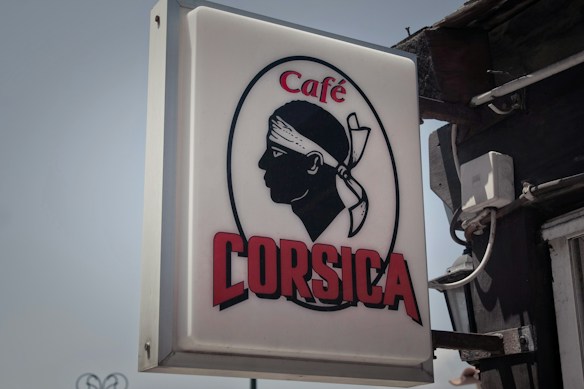 A rectangular sign displays the silhouette of a person's head with a bandana over the forehead, accompanied by the text 'Caf&eacute; Corsica'. The silhouette is in a bold black, while the text is a deep red and uppercase. The background of the sign is white, and the image is attached to a dark wall with visible wires.