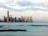 A city skyline with numerous high-rise buildings situated along the waterfront, reflecting in the calm water of the bay. The sky is partly cloudy with soft pink, blue, and gray hues.