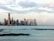 A city skyline with numerous high-rise buildings situated along the waterfront, reflecting in the calm water of the bay. The sky is partly cloudy with soft pink, blue, and gray hues.