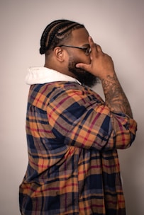A person in profile view is seen with braided hair and a well-groomed beard, wearing glasses. The individual is dressed in a colorful plaid shirt layered over a white hoodie, and has an arm tattoo. The person poses with one hand gesturing on their face against a neutral background.