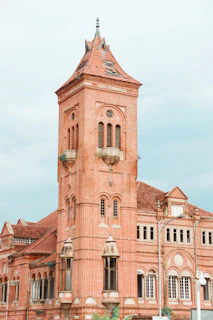 A historic red brick building with tall arched windows and a steeply pitched roof. The structure features decorative elements and a tower with a pointed top.