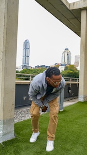 A man wearing a gray hoodie and beige cargo pants is leaning forward slightly under a concrete structure. He appears to be smiling or laughing, with glasses on. The background features an urban cityscape with tall buildings and lush green trees.