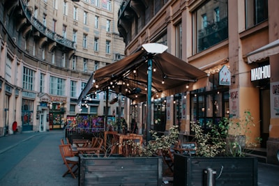 An outdoor cafe with wooden tables and chairs under a large brown canopy is set in a curved, historic street. String lights decorate the canopy, and there are potted plants along the seating area. Surrounding buildings feature ornate architecture with multiple windows.