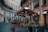 An outdoor cafe with wooden tables and chairs under a large brown canopy is set in a curved, historic street. String lights decorate the canopy, and there are potted plants along the seating area. Surrounding buildings feature ornate architecture with multiple windows.