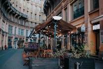 An outdoor cafe with wooden tables and chairs under a large brown canopy is set in a curved, historic street. String lights decorate the canopy, and there are potted plants along the seating area. Surrounding buildings feature ornate architecture with multiple windows.