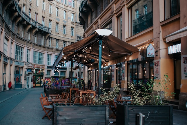 An outdoor cafe with wooden tables and chairs under a large brown canopy is set in a curved, historic street. String lights decorate the canopy, and there are potted plants along the seating area. Surrounding buildings feature ornate architecture with multiple windows.