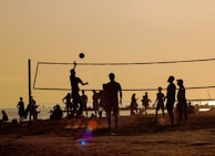 Silhouettes of people playing beach volleyball at sunset. The sun casts a warm golden hue over the scene, with the net and players sharply outlined against the bright sky. In the background, more people can be seen relaxing by the water, creating a lively and energetic atmosphere.