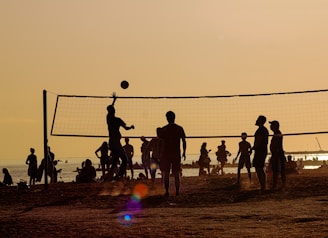 Silhouettes of people playing beach volleyball at sunset. The sun casts a warm golden hue over the scene, with the net and players sharply outlined against the bright sky. In the background, more people can be seen relaxing by the water, creating a lively and energetic atmosphere.