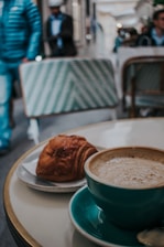 A cup of coffee in a teal cup sits on a round table next to a croissant on a small plate. In the background, there are blurred figures walking and seated chairs with a green and white pattern. It seems to be a café setting with a relaxed atmosphere.
