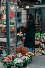 A person with long hair stands in front of a flower shop, holding a red bag and looking at various floral arrangements. Several potted flowers and bouquets are displayed outside the shop, with a variety of colorful blossoms including sunflowers, roses, and other mixed flowers. The setting appears to be urban, with buildings and lighting visible in the background.