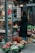 A person with long hair stands in front of a flower shop, holding a red bag and looking at various floral arrangements. Several potted flowers and bouquets are displayed outside the shop, with a variety of colorful blossoms including sunflowers, roses, and other mixed flowers. The setting appears to be urban, with buildings and lighting visible in the background.
