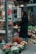 A person with long hair stands in front of a flower shop, holding a red bag and looking at various floral arrangements. Several potted flowers and bouquets are displayed outside the shop, with a variety of colorful blossoms including sunflowers, roses, and other mixed flowers. The setting appears to be urban, with buildings and lighting visible in the background.