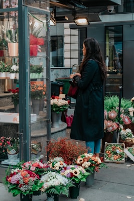 A person with long hair stands in front of a flower shop, holding a red bag and looking at various floral arrangements. Several potted flowers and bouquets are displayed outside the shop, with a variety of colorful blossoms including sunflowers, roses, and other mixed flowers. The setting appears to be urban, with buildings and lighting visible in the background.
