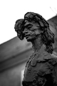 A black and white photograph of a detailed sculpture featuring a human figure with an abstract, textured surface. The sculpture appears to have a contemplative facial expression, and the background includes an out-of-focus building with a sloping roof.