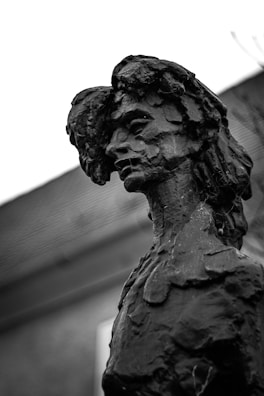 A black and white photograph of a detailed sculpture featuring a human figure with an abstract, textured surface. The sculpture appears to have a contemplative facial expression, and the background includes an out-of-focus building with a sloping roof.