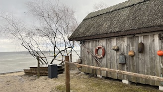 A rustic wooden cabin with a thatched roof stands near a calm beach. Various nautical items, including a life preserver, buoys, and ropes, are hung on the cabin. A leafless tree stands nearby, and the sea is visible in the background under a cloudy sky.