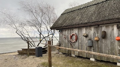 A rustic wooden cabin with a thatched roof stands near a calm beach. Various nautical items, including a life preserver, buoys, and ropes, are hung on the cabin. A leafless tree stands nearby, and the sea is visible in the background under a cloudy sky.
