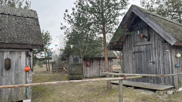 Several rustic wooden cabins are surrounded by trees and a grassy area. The cabins have thatched roofs and are decorated with various buoys and life rings. There is a simple wooden fence in the foreground, and colorful triangular flags are strung between trees.