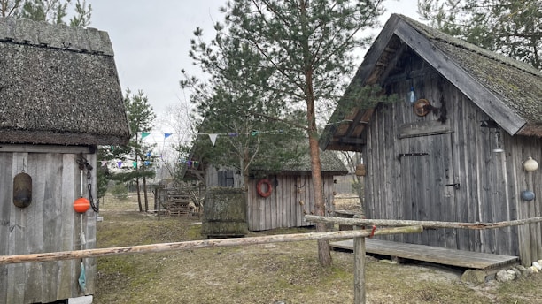 Several rustic wooden cabins are surrounded by trees and a grassy area. The cabins have thatched roofs and are decorated with various buoys and life rings. There is a simple wooden fence in the foreground, and colorful triangular flags are strung between trees.