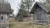 Several rustic wooden cabins are surrounded by trees and a grassy area. The cabins have thatched roofs and are decorated with various buoys and life rings. There is a simple wooden fence in the foreground, and colorful triangular flags are strung between trees.