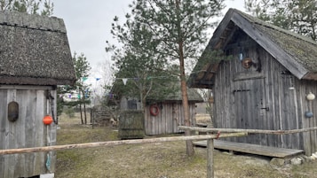 Several rustic wooden cabins are surrounded by trees and a grassy area. The cabins have thatched roofs and are decorated with various buoys and life rings. There is a simple wooden fence in the foreground, and colorful triangular flags are strung between trees.