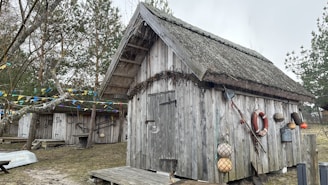 A rustic wooden cabin with a thatched roof is set in a natural environment surrounded by trees. The exterior is adorned with nautical-themed decorations, including a life preserver and fishing nets. Colorful triangular flags are strung across the scene, adding a festive touch. The ground appears to be covered with light grass and patches of sand.
