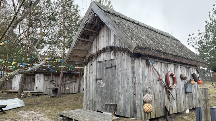 A rustic wooden cabin with a thatched roof is set in a natural environment surrounded by trees. The exterior is adorned with nautical-themed decorations, including a life preserver and fishing nets. Colorful triangular flags are strung across the scene, adding a festive touch. The ground appears to be covered with light grass and patches of sand.
