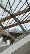 A modern architectural structure featuring glass panels and wooden beams provides a view of a distant city skyline. The urban landscape consists of various skyscrapers set against a cloudy sky, with a mix of greenery and buildings in the foreground.