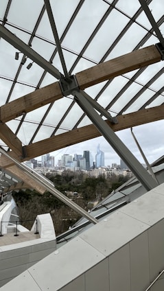 A modern architectural structure featuring glass panels and wooden beams provides a view of a distant city skyline. The urban landscape consists of various skyscrapers set against a cloudy sky, with a mix of greenery and buildings in the foreground.