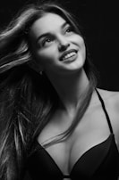 A black and white portrait of a woman with long hair, wearing a bra. She is looking upwards with a gentle smile and her hair flows softly, creating a dynamic and graceful appearance.