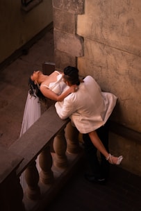 A man in a white suit is romantically holding a woman in a white dress as she leans back over a stone balustrade. The scene is captured from an elevated angle, emphasizing the intimacy and drama of the setting. The sun casts warm shadows on the couple and the surrounding stone architecture.