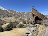 A wooden mining structure is perched on a rocky hilltop, overlooking a small town situated in a valley. The town is nestled among arid, mountainous terrain with patches of snow on the peaks. Sparse vegetation with scattered trees covers the slopes.