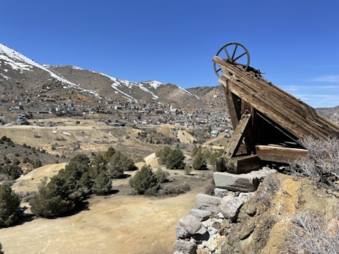 A wooden mining structure is perched on a rocky hilltop, overlooking a small town situated in a valley. The town is nestled among arid, mountainous terrain with patches of snow on the peaks. Sparse vegetation with scattered trees covers the slopes.