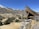A wooden mining structure is perched on a rocky hilltop, overlooking a small town situated in a valley. The town is nestled among arid, mountainous terrain with patches of snow on the peaks. Sparse vegetation with scattered trees covers the slopes.