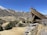 A wooden mining structure is perched on a rocky hilltop, overlooking a small town situated in a valley. The town is nestled among arid, mountainous terrain with patches of snow on the peaks. Sparse vegetation with scattered trees covers the slopes.