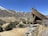 A wooden mining structure is perched on a rocky hilltop, overlooking a small town situated in a valley. The town is nestled among arid, mountainous terrain with patches of snow on the peaks. Sparse vegetation with scattered trees covers the slopes.