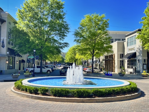 A vibrant urban scene with a circular fountain at the center, surrounded by manicured greenery and brick paving. Tall, lush trees line the street, flanking modern commercial buildings that house shops and cafes. Several parked cars are visible along the street, adding to the bustling yet serene atmosphere.
