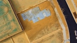 An aerial view of a construction site with a foundation layout. The site is surrounded by areas of dirt and soil, with a paved road running along one side.