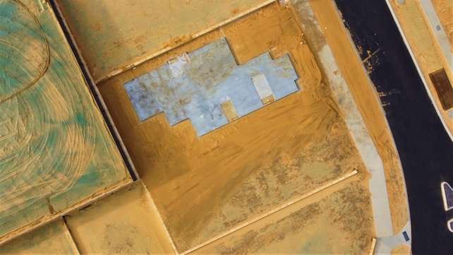 An aerial view of a construction site with a foundation layout. The site is surrounded by areas of dirt and soil, with a paved road running along one side.