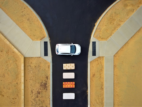 Aerial view of a white car parked at a T-intersection on a dark asphalt road. Three mattresses are lined up vertically on the road behind the car. The surrounding area consists of sandy terrain with concrete sidewalks.