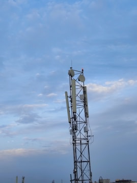 A tall communication tower stands against a backdrop of a partly cloudy sky. The tower is equipped with multiple antennas and dishes, with some wiring visible around its structure. The lower part of the image includes a faint view of other distant structures and rooftops, creating an urban or industrial setting.