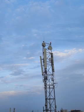A tall communication tower stands against a backdrop of a partly cloudy sky. The tower is equipped with multiple antennas and dishes, with some wiring visible around its structure. The lower part of the image includes a faint view of other distant structures and rooftops, creating an urban or industrial setting.
