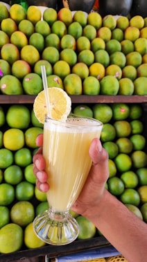A hand holding a glass of lemon juice with a slice of lemon on the rim. The background is filled with an arrangement of green and slightly yellow limes.