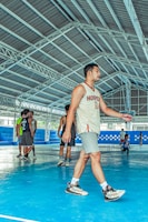 Several young men are on an indoor basketball court, one wearing a jersey with the number 10 and the word 'Hooper'. The court features a blue floor and is surrounded by a metal framework structure. Other individuals in jerseys are seen in the background.