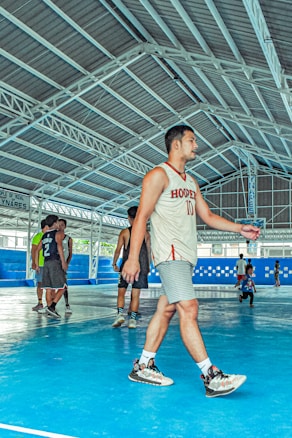 Several young men are on an indoor basketball court, one wearing a jersey with the number 10 and the word 'Hooper'. The court features a blue floor and is surrounded by a metal framework structure. Other individuals in jerseys are seen in the background.