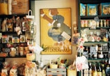 A small store interior filled with various packaged goods displayed on dark shelves. A prominent vintage poster with the words 'Champagne Kupferberg Gold' is centered on the wall. The shelves are stocked with snacks, pasta, condiments, and decorative items, many labeled with tags. Brightly colored baskets and gift bags are arranged at the bottom.