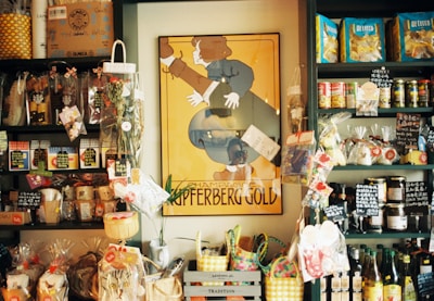 A small store interior filled with various packaged goods displayed on dark shelves. A prominent vintage poster with the words 'Champagne Kupferberg Gold' is centered on the wall. The shelves are stocked with snacks, pasta, condiments, and decorative items, many labeled with tags. Brightly colored baskets and gift bags are arranged at the bottom.